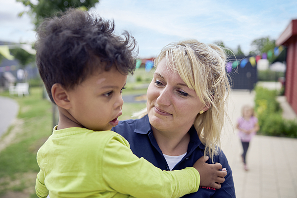 Erzieherin hält ein Kind auf dem Arm. Sie sind im Garten.