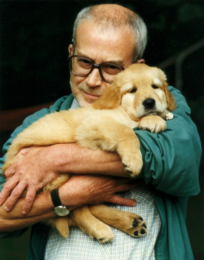 Ein älterer Mann mit Brille hält liebevoll einen Golden Retriever-Welpen im Arm. Beide wirken ruhig und vertraut.