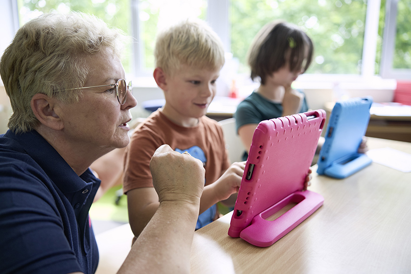 Eine Schulbegleiterin unterstützt ein Grundschulkind im Unterricht bei der Nutzung eines Tablets im Klassenzimmer.