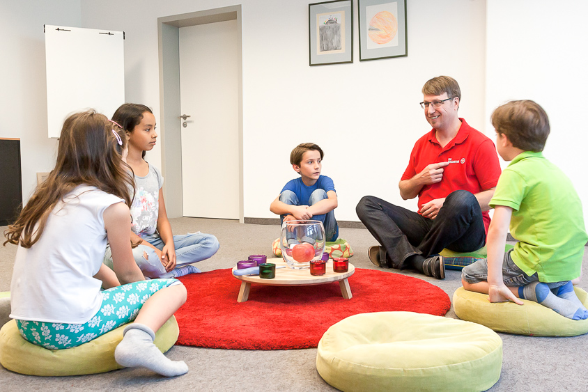 Kinder und ein Betreuer sitzen auf Kissen um einen kleinen Tisch mit Kerzen und Dekoration.
