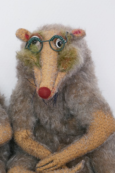 Eine graue Ratten-Handpuppe mit roter Nase und grüner Brille sitzt vor einem hellen Hintergrund.