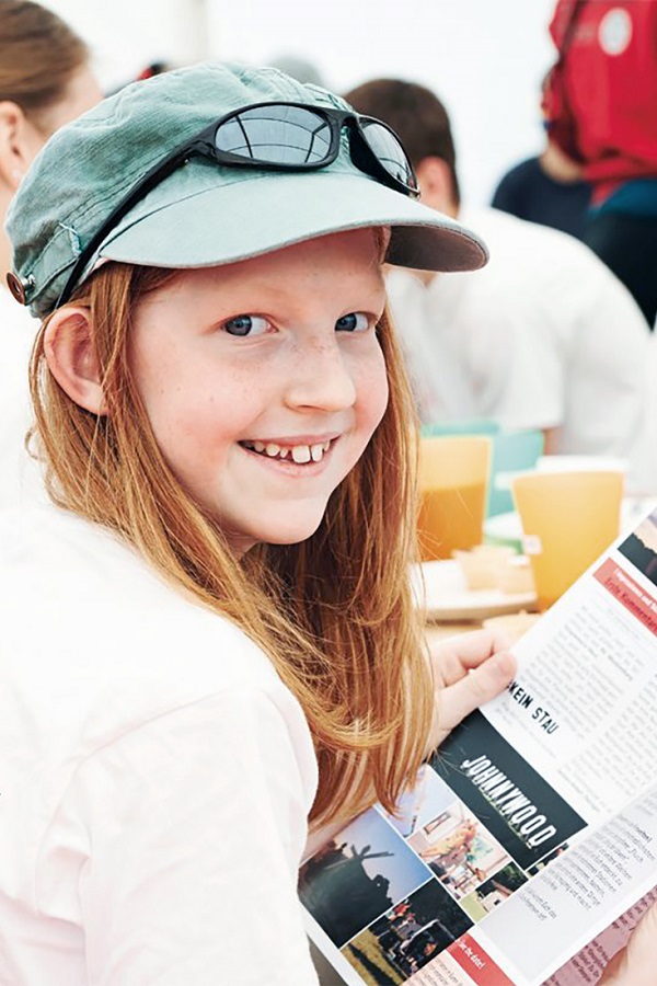 Ein Mädchen mit Kappe und Sonnenbrille liest lächelnd eine Broschüre.
