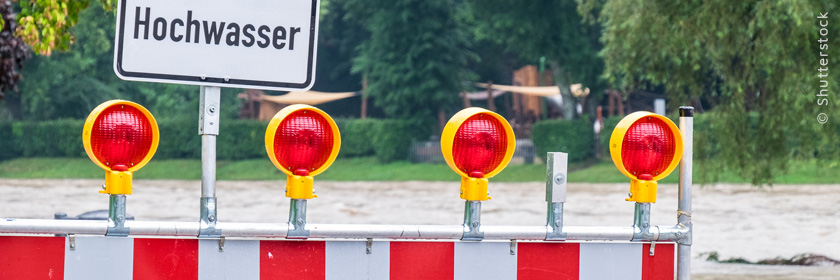 Absperrung mit rot-weißen Warnbaken und Blinklichtern vor einem Hochwassergebiet.