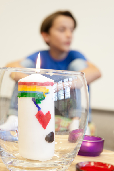 Brennende Kerze mit Regenbogen und Herz im Glas, unscharf sitzendes Kind im Hintergrund