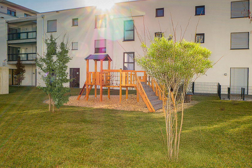 Grüner Außenbereich einer Kita, der von Wohnhäusern umgeben ist. In der Mitte ist ein großer Sandbereich auf welchem ein Klettergerüst aus Holz steht.