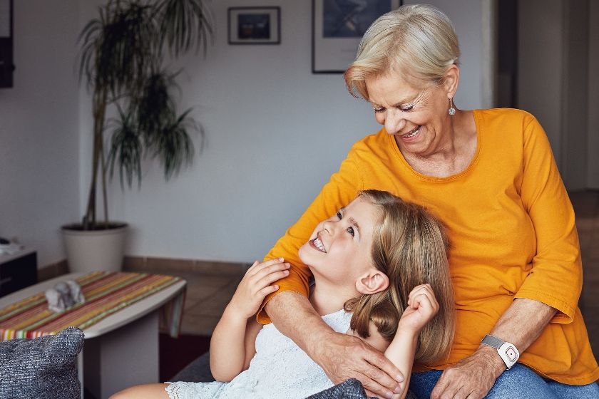 Eine ältere Frau in gelbem Pullover sitzt lächelnd auf dem Sofa und umarmt liebevoll ein kleines Mädchen, das sie fröhlich anschaut.