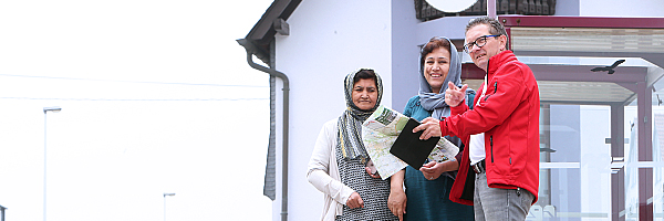 Ein Mitarbeiter der Flüchtlingshilfe an einer Bushaltestelle zeigt zwei Frauen mit Kopftuch den Weg.