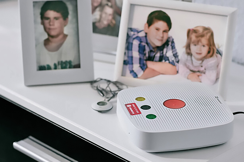 Hausnotrufgerät der Johanniter Hanau auf einem weißen Tisch. Im Hintergrund Bilder einer Familie.
