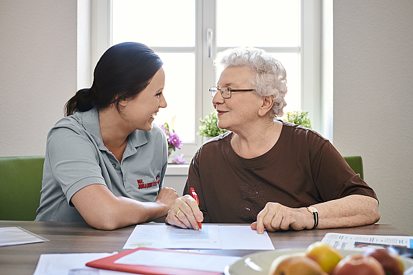 Eine ältere Frau und eine Pflegekraft sitzen zusammen an einem Tisch und blicken sich an