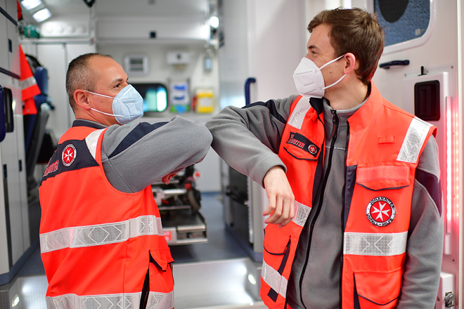 Zwei Mitarbeiter mit Johanniter-Weste tragen Maske und begrüßen sich durch Berührung der Ellenbogen