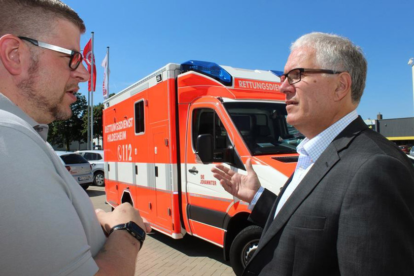 Maik Hüpper unterhält sich mit Ottmar von Holtz. Im Hintergrund steht ein Einsatzfahrzeug der Johanniter. 