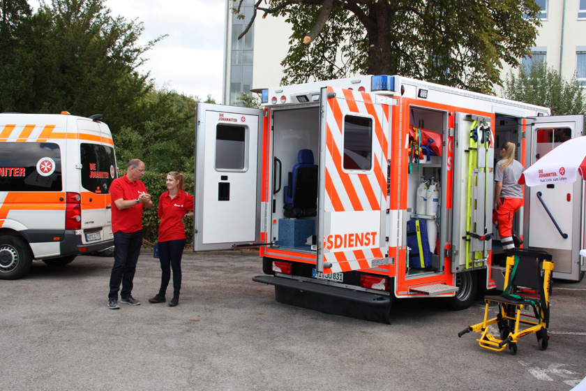 Johanniter Mitarbeitende und ein Rettungswagen mit geöffneten Seiten- und Hecktüren zum Besichtigen.