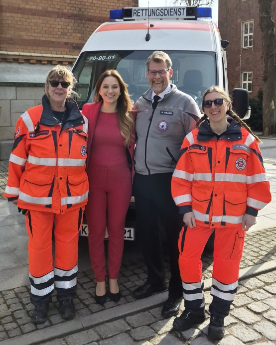 Eine Gruppe von vier Personen steht vor einem Rettungswagen und blicken in die Kamera.