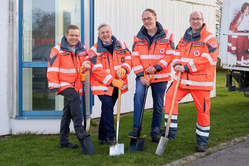 Vier Männer in Johanniter-Einsatzjacken setzen jeweils einen Spaten auf einer Wiese vor einem Gebäude an.