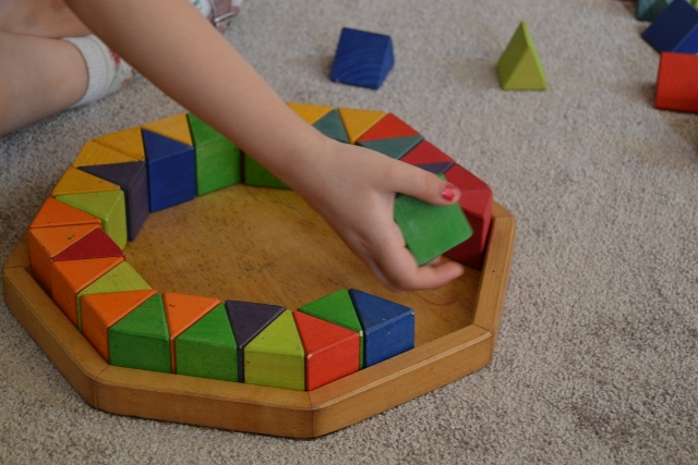 Eine Kinderhand setzt einen grünen Baustein in ein Muster aus anderen Holzbausteinen ein.