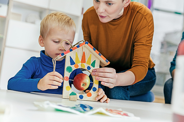 Ein Junge im Kindergarten baut mit seiner Erzieherin ein Vogelhäuschen mit einem Apfel.