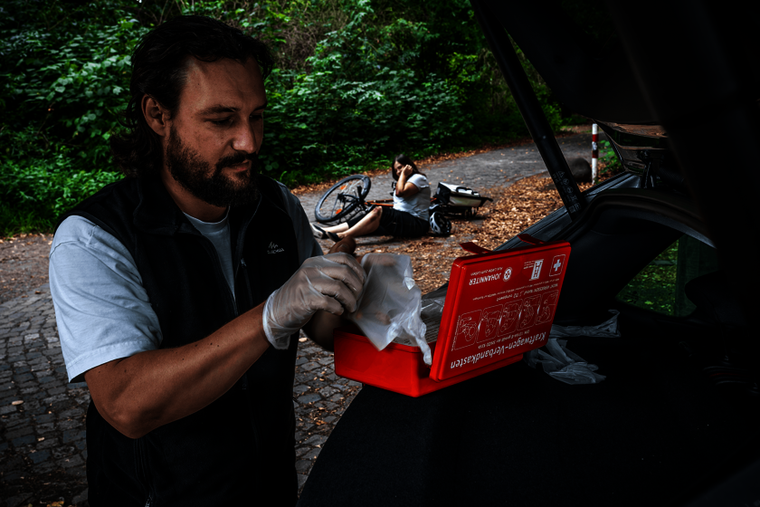 Ein Mann am geöffneten Kofferraum holt Verbandsmaterial aus einem Erste-Hilfe-Kasten, im Hintergrund sitzt eine gestürzte Radfahrerin am Boden.