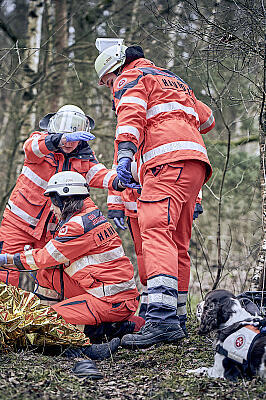 Drei Mitglieder der Rettungshundestaffel versorgen einen gerade durch den Rettungshund gefunden Mann im Wald.