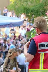 Drei ehrenamtliche Sanitätshelfende beobachten von einer Erhöhung eine Menschenmenge auf einem Stadtfest. Sie tragen Einsatzkleidung und Rucksack.