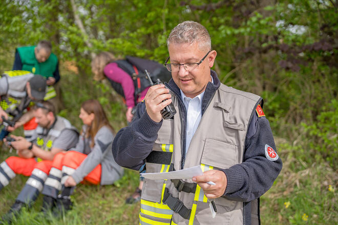 Ein Johanniter steht vor grünen Bäumen und spricht in ein Walkie-Talkie und liest dabei von einem Zettel ab.