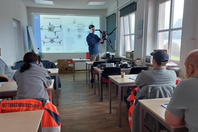 Zu sehen sind vier Personen, die am Unterricht teilnehmen. Vorne steht eine Person, die erklärt, wie eine Drohne funktioniert.