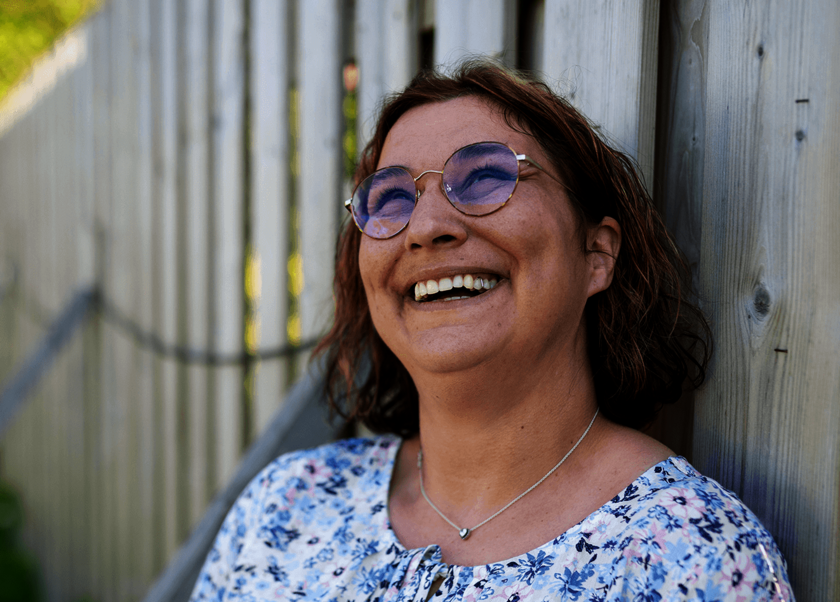 Nahaufnahme: Pflegerin lehnt an einem Holzzaun und lacht, sonniger Hintergrund.  