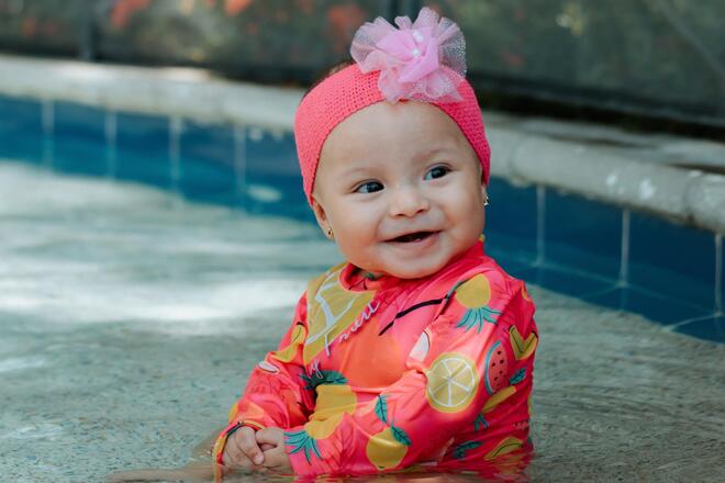 Fröhliches Baby mit bunter Badebekleidung und Stirnband sitzt in einem hellblauen gekachelten Pool