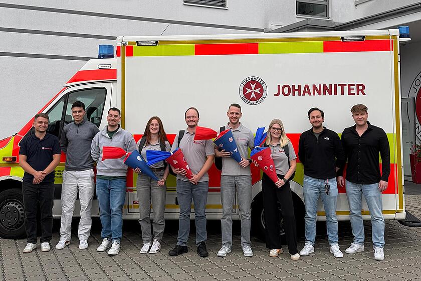 Die neuen Mitarbeitenden im Johanniter-Rettungsdienst sind bereit für ihren Einsatz. Gemeinsam stehen sie in einer Gruppe zusammen und lächeln in die Kamera. Im Hintergrund ist ein Johanniter - Rettungswagen zu sehen.