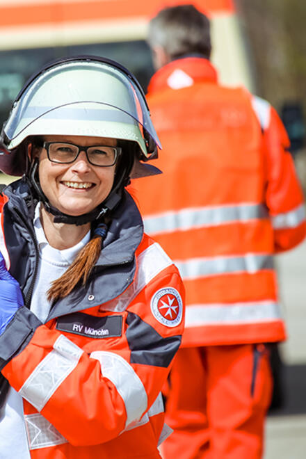 Eine lächelnde Johanniter-Helferin in leuchtroter Einsatzkleidung und Schutzhelm trägt einen roten Rettungsrucksack. Im Hintergrund ist ein weiteres Teammitglied und ein Einsatzfahrzeug zu sehen.