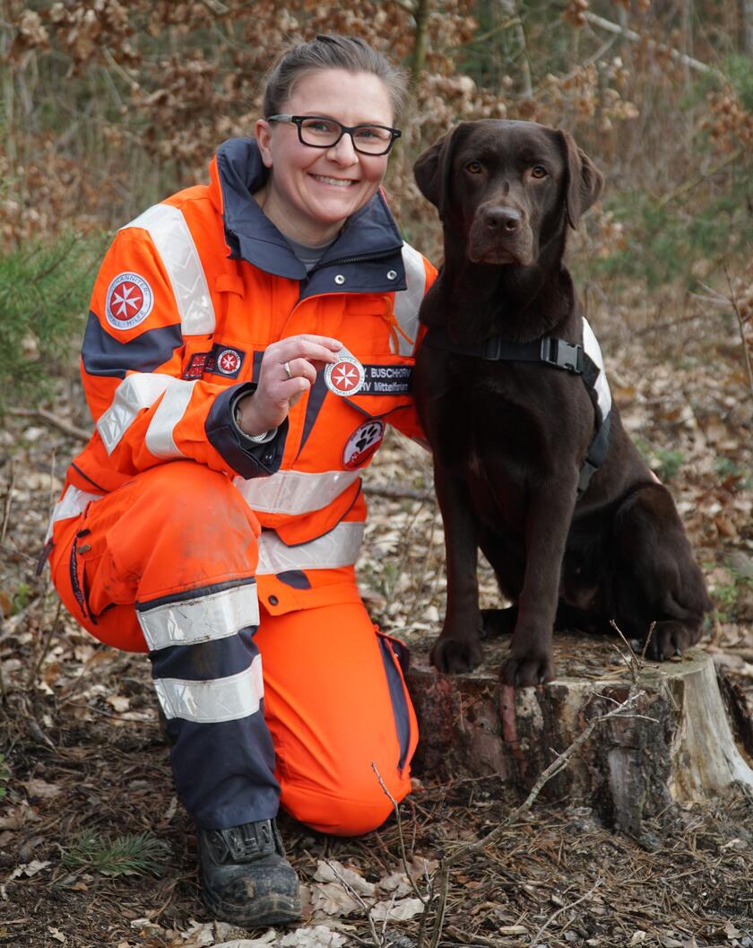 Eine Frau in Einsatzkleidung der Johanniter-Unfall-Hilfe kniet im Wald auf dem Boden und lächelt in die Kamera. Neben ihr sitzt ein brauner Labrador-Retriever mit Geschirr auf einem Baumstumpf. Beide wirken aufmerksam und freundlich. Die Frau trägt eine Brille und hat ihre Haare zum Zopf gebunden. Das Umfeld ist herbstlich mit braunem Laub auf dem Boden und Bäumen im Hintergrund.