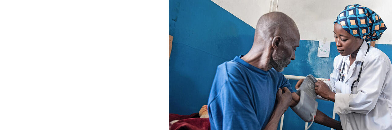 Ärztin mit Kopftuch misst Blutdruck bei einem Patienten in einem dürftig ausgestatteten Zimmer.