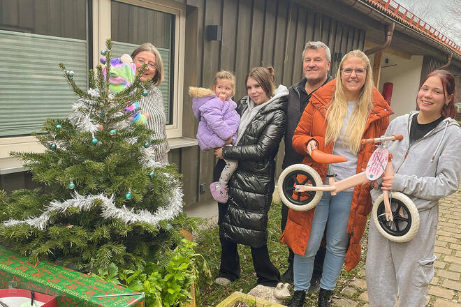 Mehrere Leute stehen um einen Weihnachtsbaum. Unter dem Baum stehen Geschenke und zwei Frauen halten ein Laufrad in der Hand.