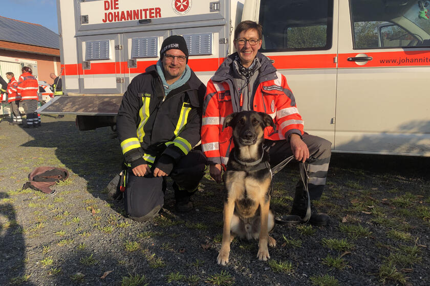 Rettungshund Poirot und Rettungshundeteam nach dem Einsatz. Im Hintergrund ist ein Einsatzfahrzeug der Johanniter-Rettungshundestaffel.  