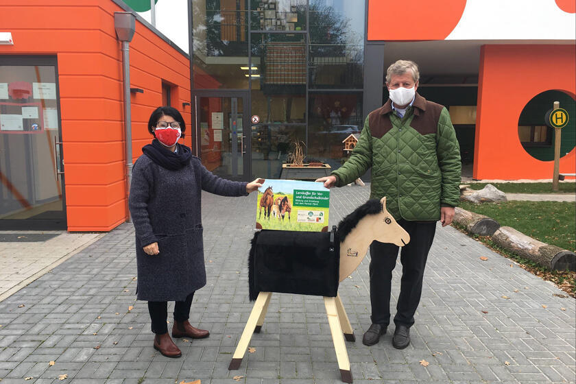Kitaleiterin und Carl Garf von Hardenberg stehen vor der Kita Elementum und zeigen das Holzpferd inklusive Lernkoffer. 