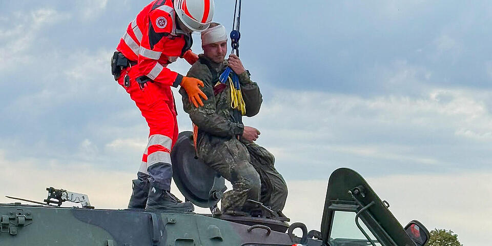 Ein Soldat mit bandagiertem Kopf wird mittels eines Krahns aus einem Truppentransportpanzer, der mit einem PKW kollidiert ist, gezogen. Ein Mann mit Einsatzkleidung und Helm steht auf dem Panzer und unterstützt.  Eine weitere Person in Einsatzkleidung hilft vom Boden aus.