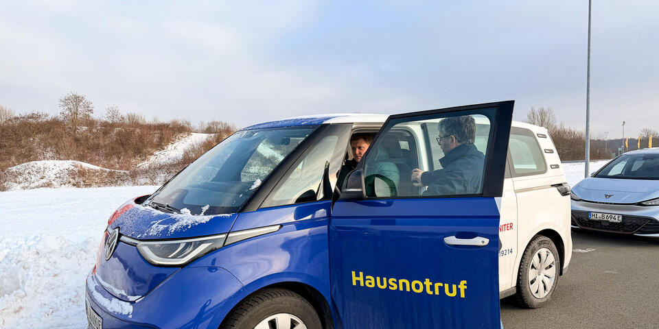 Ein Mann steht neben einem VW-Bulli. Die Fahrertür ist geöffnet und ein Fahrer sitzt im Fahrzeug