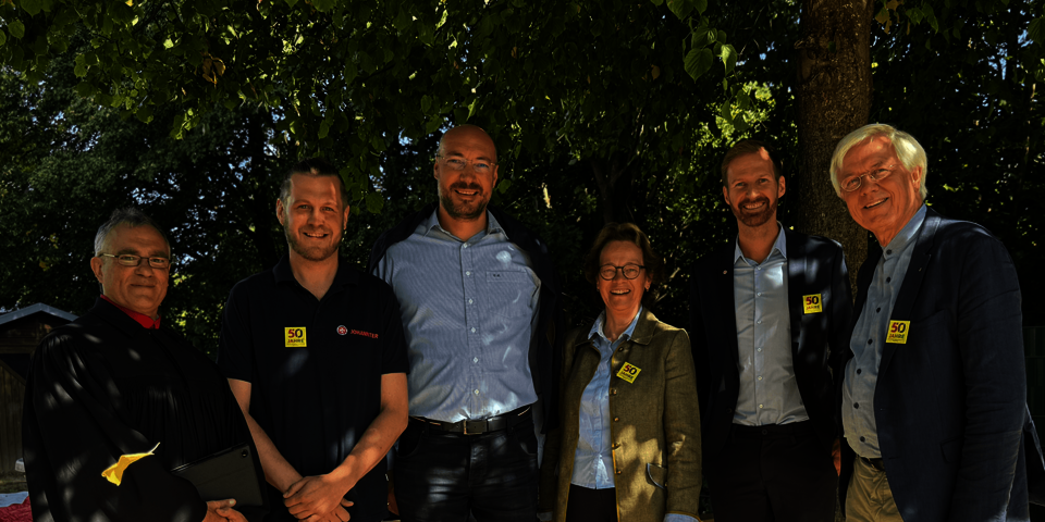 Sechs Erwachsene, darunter ein Geistlicher, stehen lachend unter einem Baum.