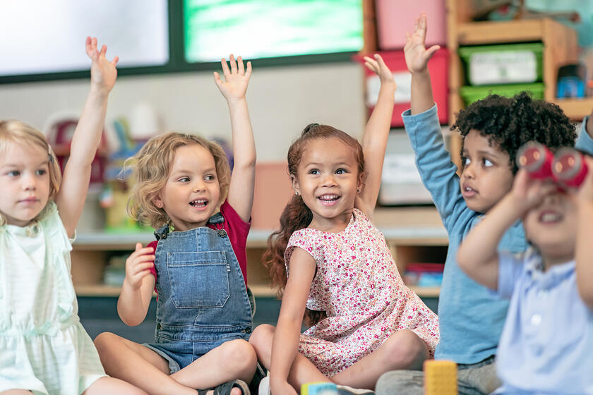 Eine Gruppe Vorschulkinder sitzt im Schneidersitz auf dem Boden und zeigt mit den Händen auf, um die Frage ihres Lehrers zu beantworten.