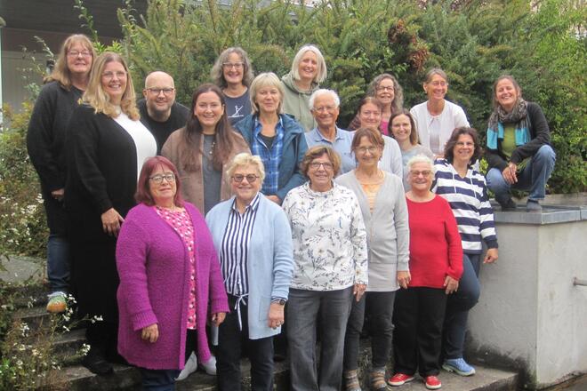 Gruppenfoto von 21 Erwachsenen, die dicht beieinander auf einer alten Steintreppe und daneben vor grünen Büschen im Freien stehen. Die Personen, überwiegend Frauen, lächeln in die Kamera.  Die freundliche Atmosphäre zeigt ein engagiertes Team, das an einem gemeinsamen Treffpunkt draußen versammelt ist.