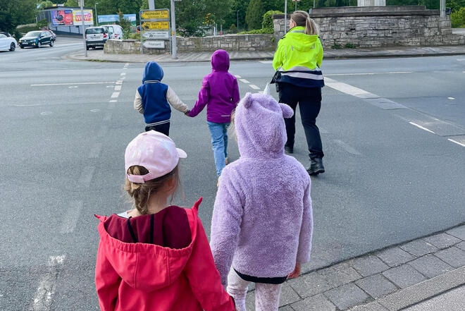 Kinder gehen in Zweipaaren Hand in Hand mit einer Polizistin über einen Fußgängerüberweg