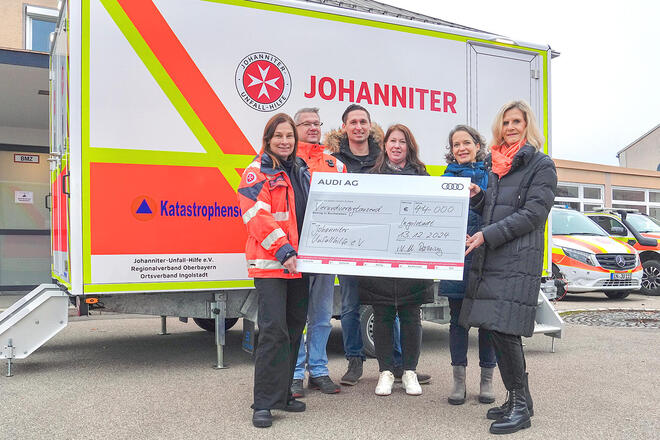Mehrere Leute stehen vor einem Johanniter Anhänger und halten einen großen Spendencheck von der Audi AG.