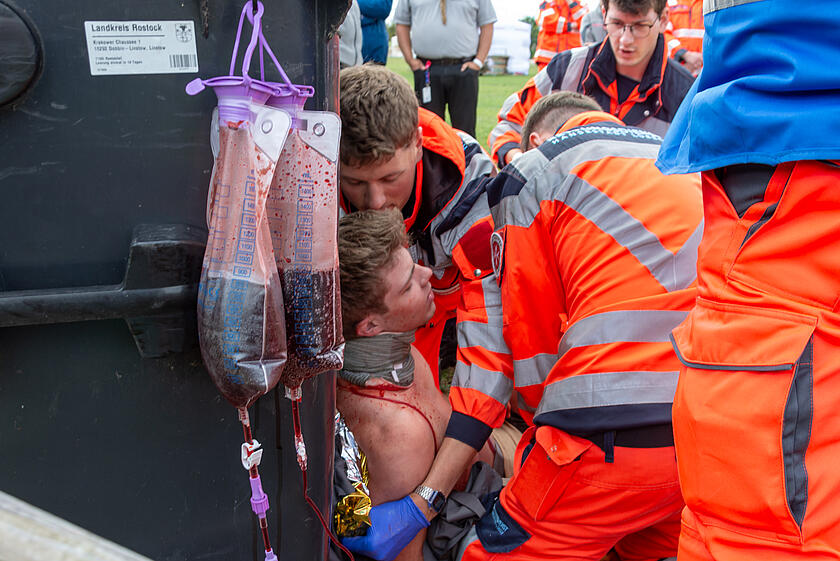 Ein schwer verletzter Mann lehnt an einer Mülltonne und wird von zwei Notfallsanitätern versorgt.