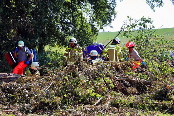 Mehrere Johanniter stehen in Mitten von hohen Sträuchern im Wald. Im Hintergrund sieht man ein aufgeschlagenes Zelt.