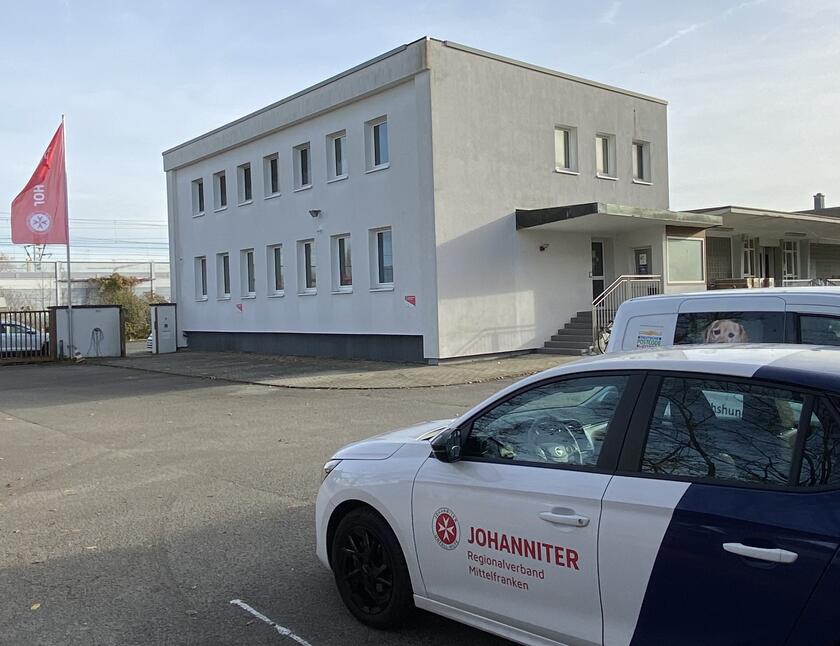 Auf dem Bild ist die Dienststelle Erlangen zusehen. Daneben eine Fahne der Johanniter und im Vordergrund ein Fahrzeug der Johanniter.