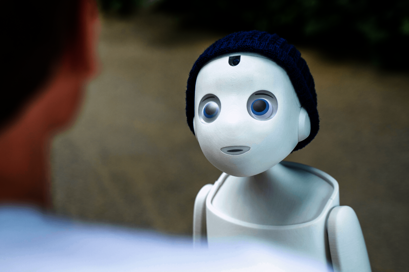 eine kleine weiße Figur mit großen Augen und blauer Mütze scheint einen Menschen zu betrachten, der ihm gegenübersteht und von dem nur ein Ausschnitt von Hals und Ohr sowie Schulter von hinten zu sehen sind.