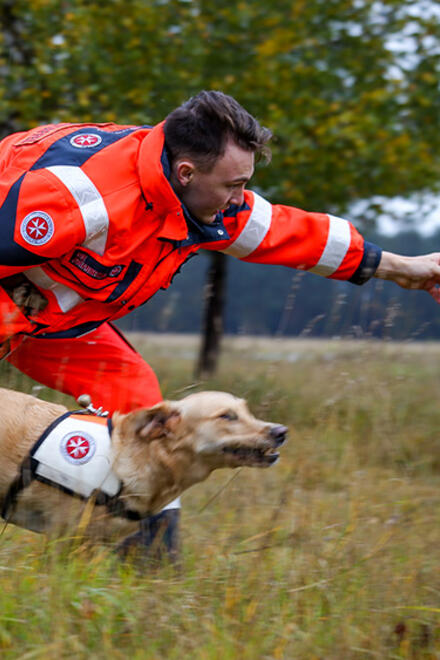 Ein Rettungshundeführer zeigt nach vorne, um seinem Hund den Suchbefehl zu geben. Der Hund, ausgestattet mit einem Johanniter-Geschirr, sprintet entschlossen über eine grasbewachsene Fläche.