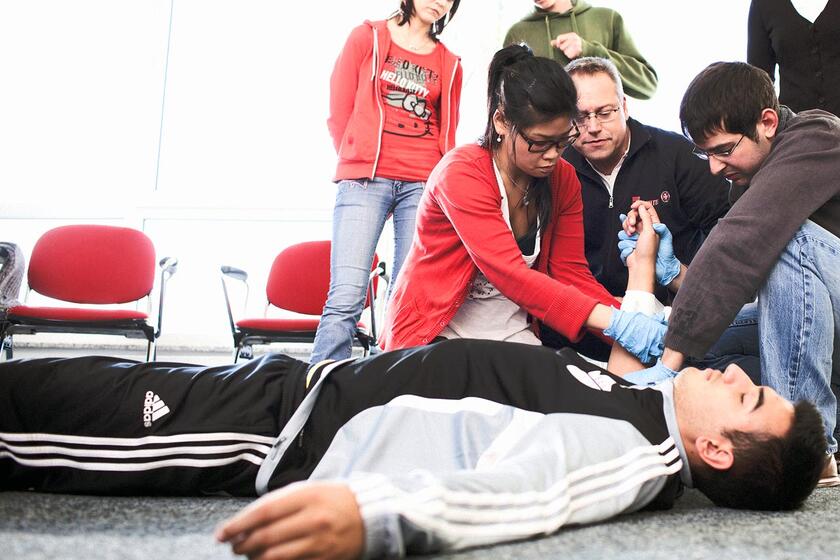 Mehrere Personen leisten bei einer Schulung gemeinsam Erste Hilfe an einem jungen Mann, der auf dem Boden liegt, und stabilisieren seinen Arm.