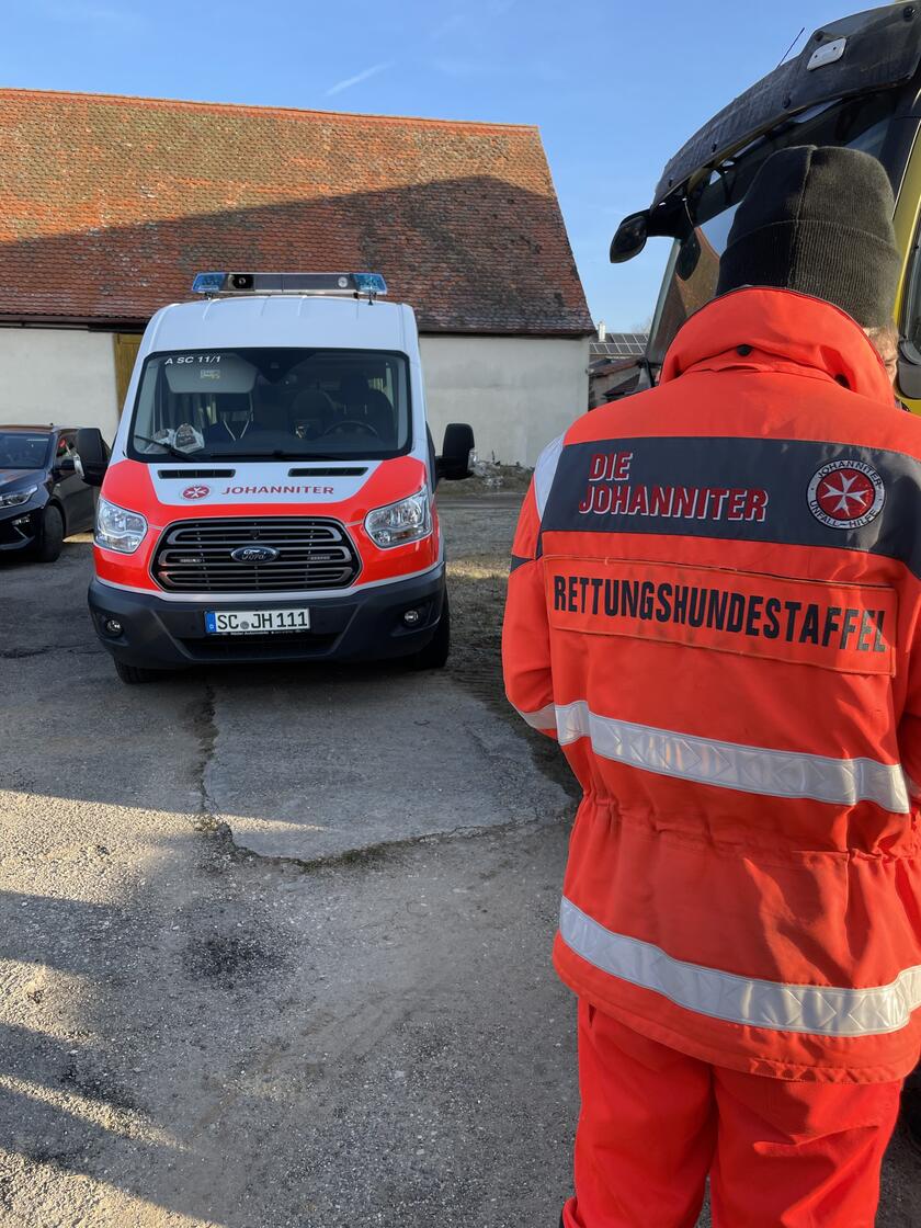 Person in Einsatzkleidung der Rettungshundestaffel steht vor einem Johanniter-Einsatzfahrzeug auf einem Hof.