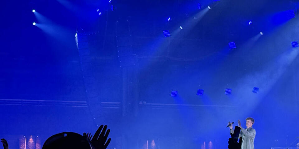 Eine große Halle, blau beleuchtet mit Zuschauern. Ein Mann mit Mikro auf einer Bühne.