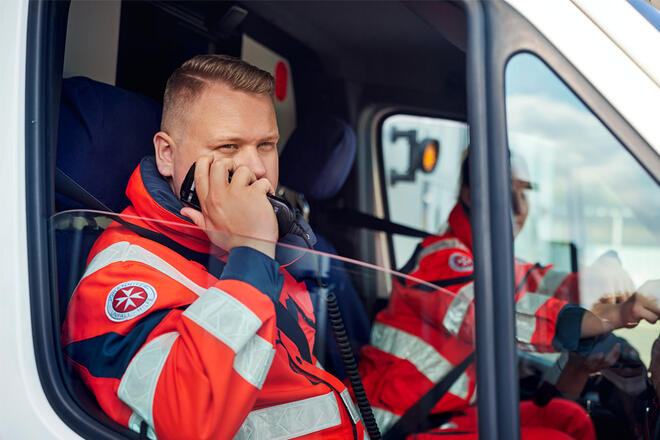 Zwei Johanniter sitzen in Einsatzkleidung in einem Einsatzfahrzeug und einer von ihnen spricht in ein Funkgerät.